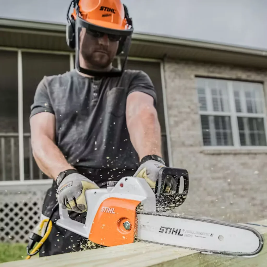 A person wearing protective gloves and a helmet operates a STIHL chainsaw to cut a piece of wood. Sawdust and wood chips scatter as the chainsaw makes contact. The background features a brick house with windows and a porch, adding a home improvement or outdoor maintenance context. The worker's protective gear emphasizes the importance of safety while using power tools.