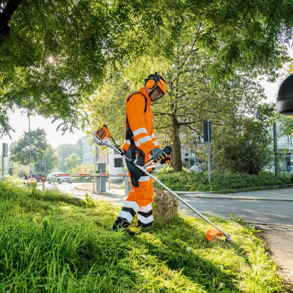 A worker wearing an orange safety suit with reflective stripes, a helmet, and ear protection operates a grass trimmer along a roadside. Lush greenery surrounds the area, with trees and bushes in the background. The street nearby has a few vehicles and buildings visible, emphasizing an urban maintenance setting. The worker’s protective gear highlights the importance of safety in outdoor labor.